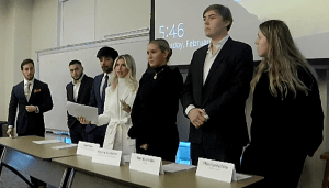 7 people standing behind table, presenting debate