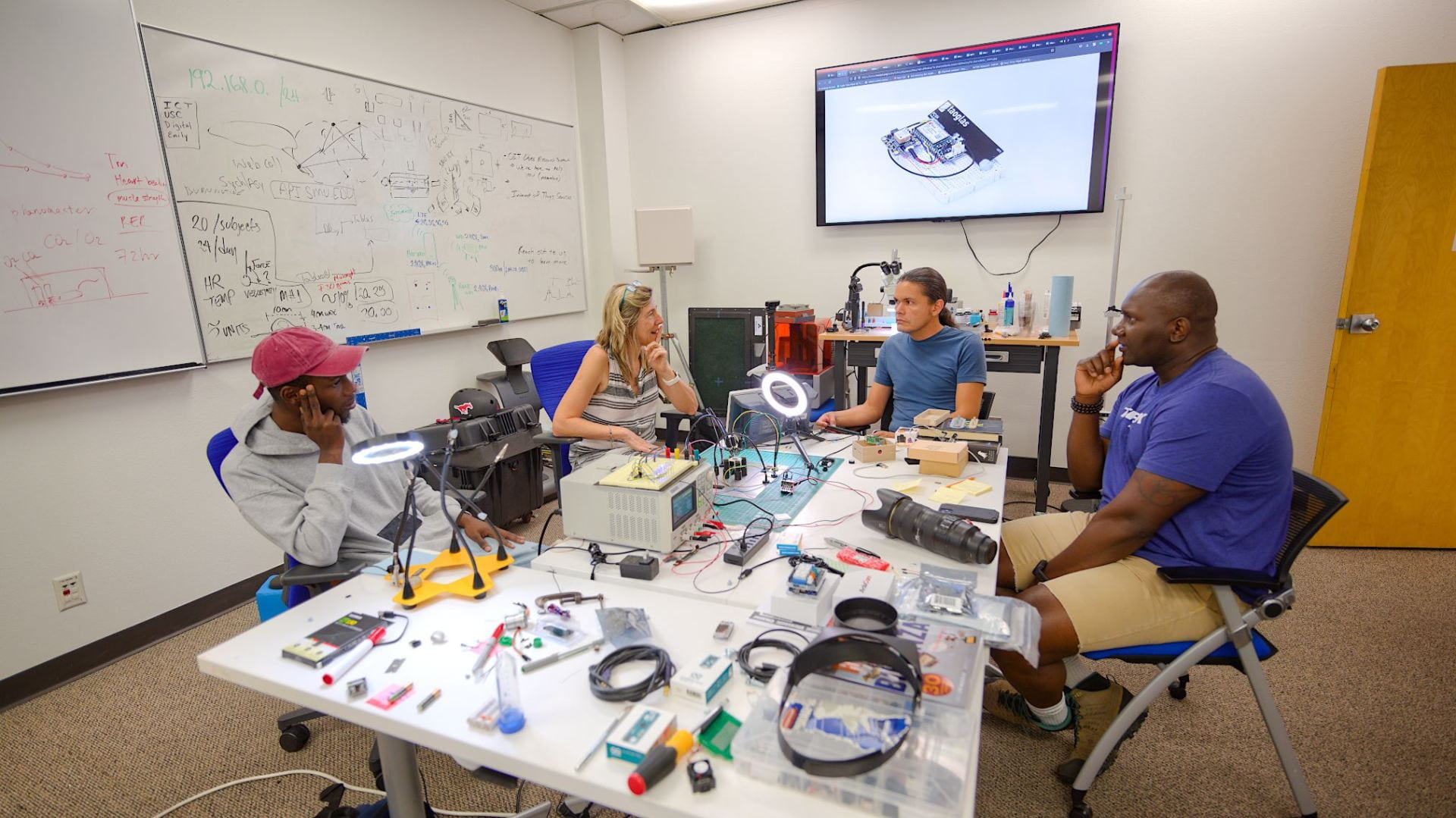 A group of people meeting in a lab at a table filled with equipment.