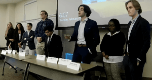 8 people standing behind table, presenting debate