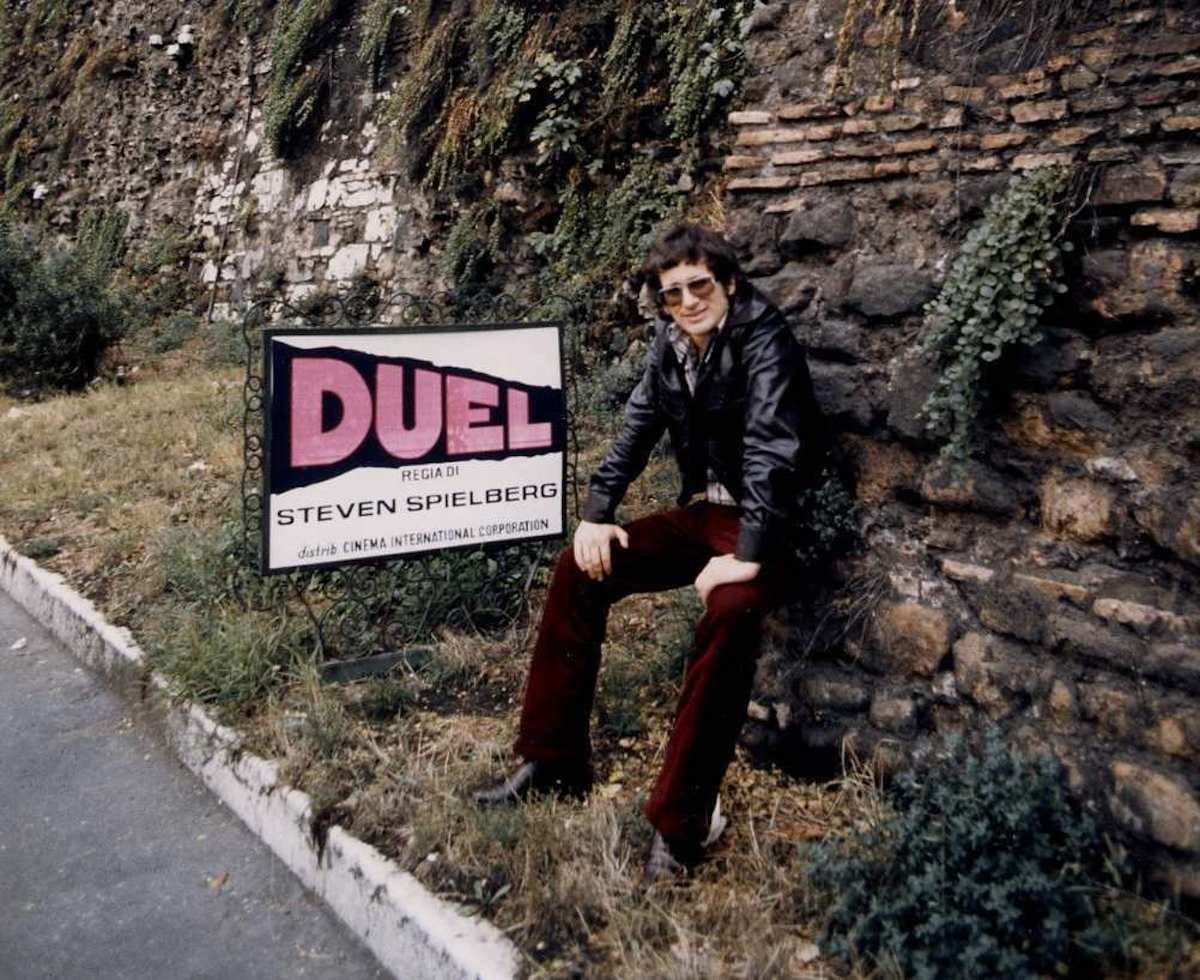 Photo of a young Steven Spielberg next to a sign advertising his film, Duel.